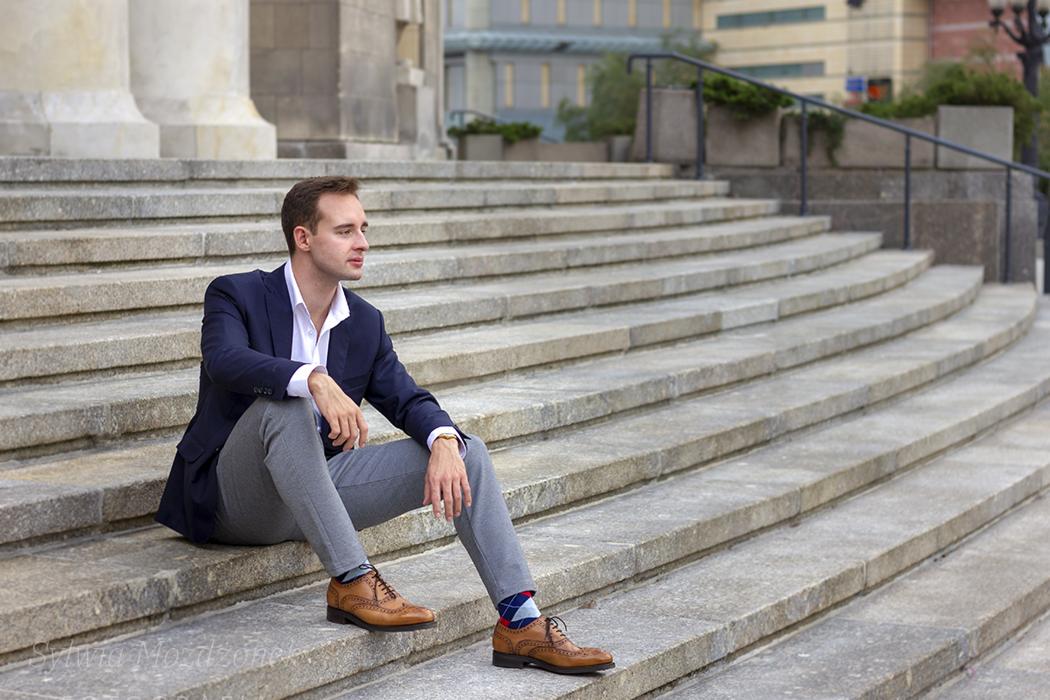 Mężczyzna w granatowej marynarce i szarych spodniach siedzi na kamiennych schodach, eksponując brązowe buty i skarpetki w romby, z budynkiem w tle.