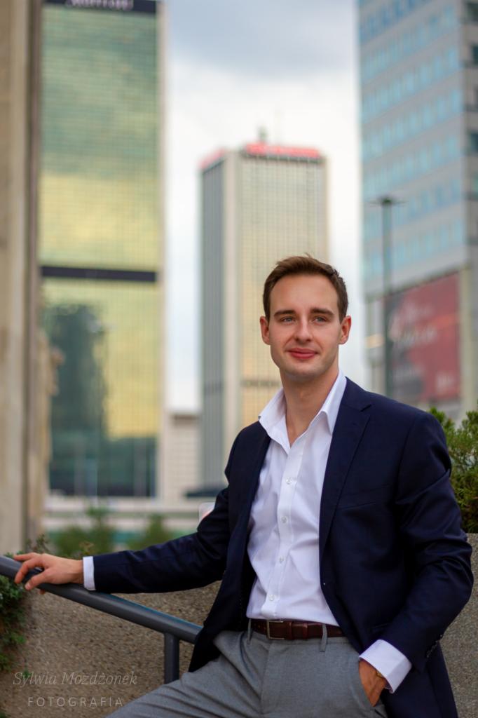 Młody mężczyzna w garniturze pozuje na tle wysokich budynków w mieście, opierając się o metalową barierkę.