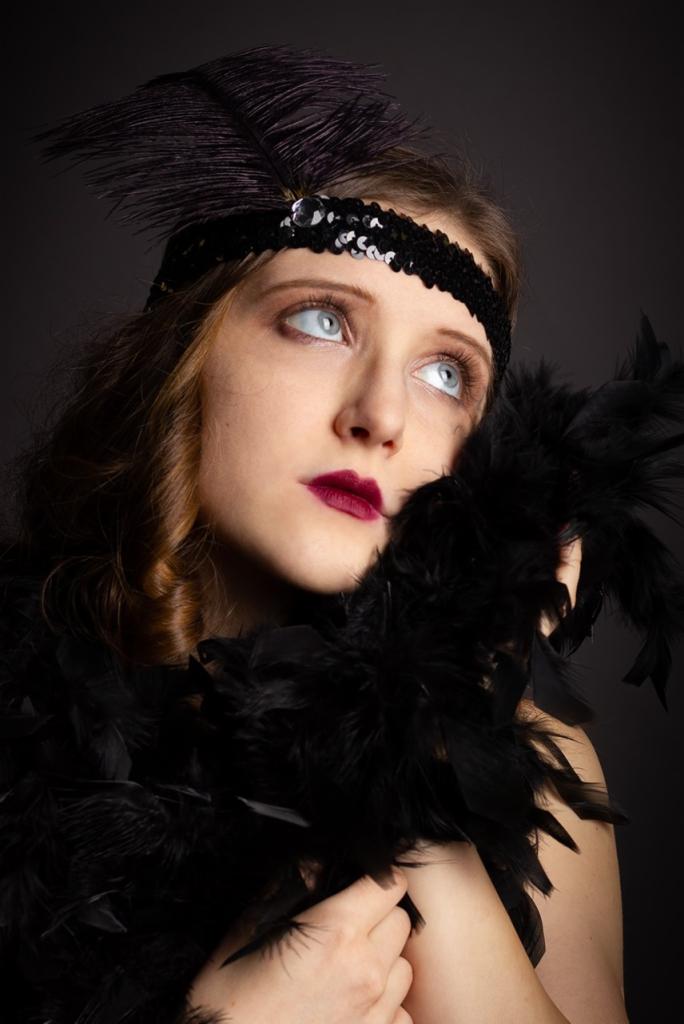 Portret kobiety w stylu lat 20. z piórami, cekinową opaską i ciemnoczerwoną szminką, spoglądającej w górę na ciemnym tle.