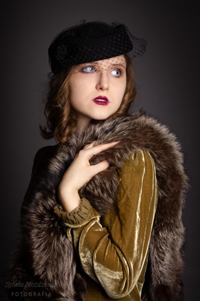 Portret kobiety w czarnym kapeluszu z woalką, futrzanym szalu i aksamitnej sukni w odcieniach złota, pozującej na ciemnoszarym tle.