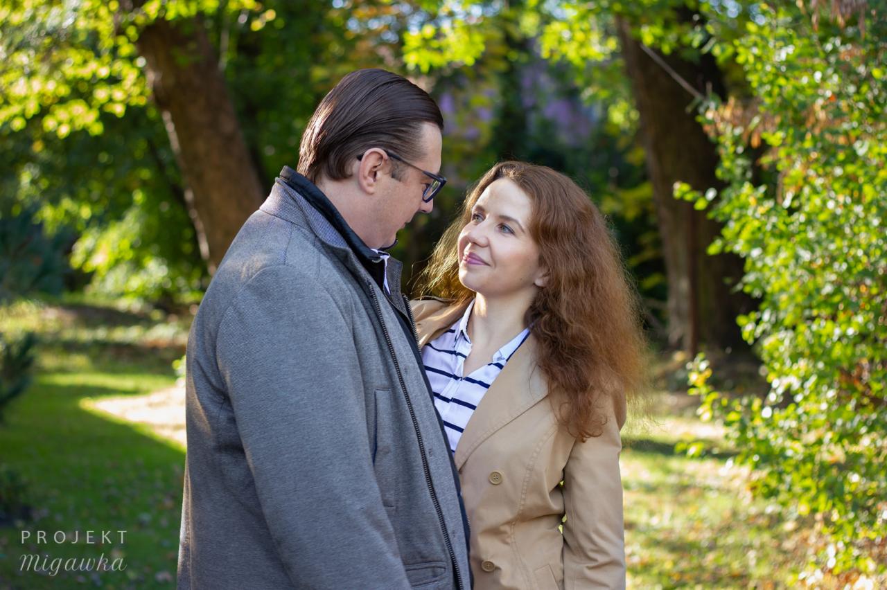 Para zakochanych, mężczyzna w okularach i szarej kurtce oraz kobieta w beżowym płaszczu, patrzą na siebie z uśmiechem w plenerze, z zielonym tłem drzew i krzewów w słoneczny dzień.