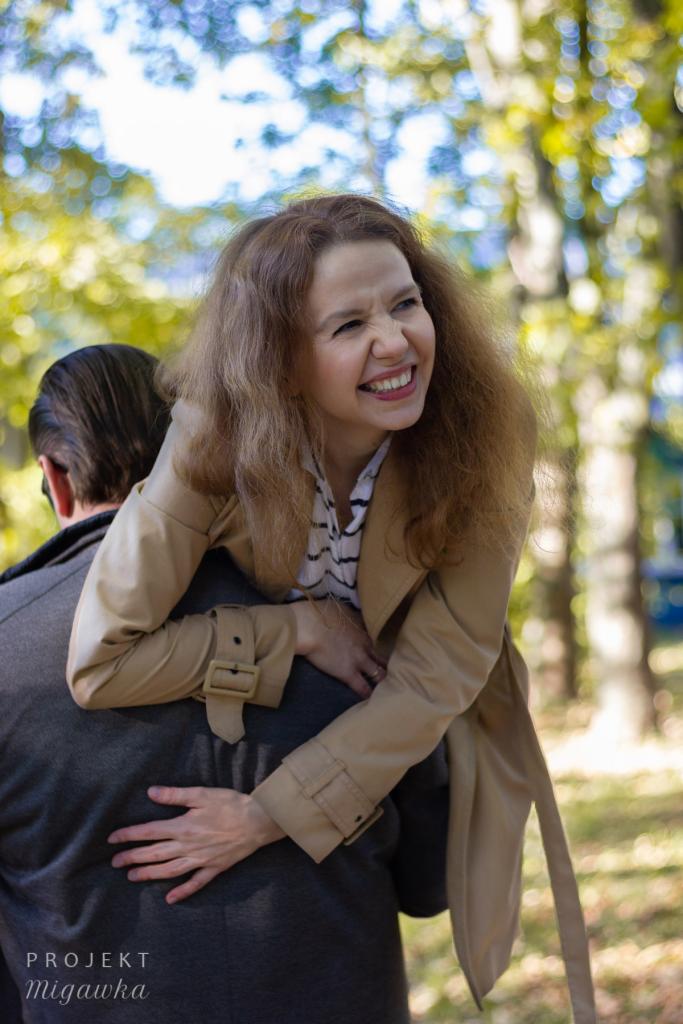 Uśmiechnięta kobieta w beżowym płaszczu, z zarzuconymi rękami na ramionach mężczyzny w szarym płaszczu, w tle rozmyte drzewa i słońce.