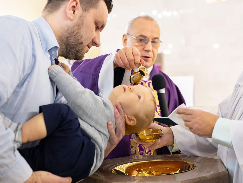 Ksiądz w fioletowej szacie polewa wodą głowę niemowlęcia trzymanego przez ojca podczas ceremonii chrztu, widoczny mikrofon i marmurowa chrzcielnica.