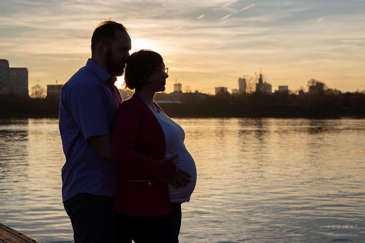 Silhouetted couple expecting a baby, standing by the river at sunset with city skyline in the background.