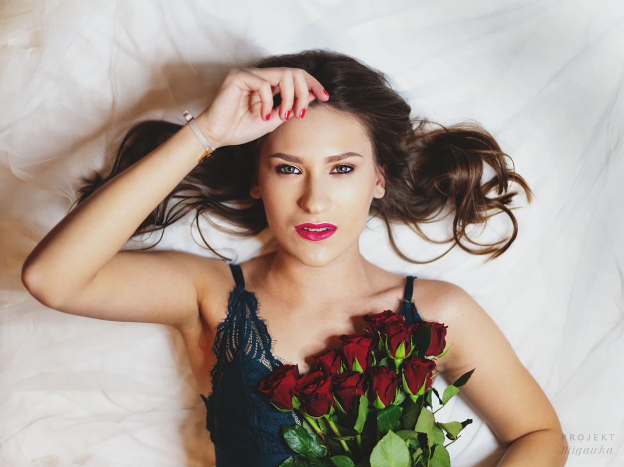 Portret kobiety leżącej na białej pościeli z bukietem czerwonych róż, ujęcie z góry, delikatny makijaż i naturalne światło.