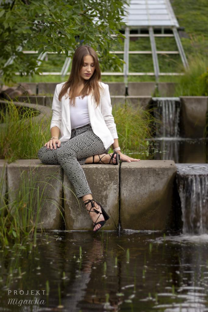 Młoda kobieta w białym żakiecie i wzorzystych spodniach siedzi na betonowym murku przy kaskadzie wodnej, pozując do sesji zdjęciowej.
