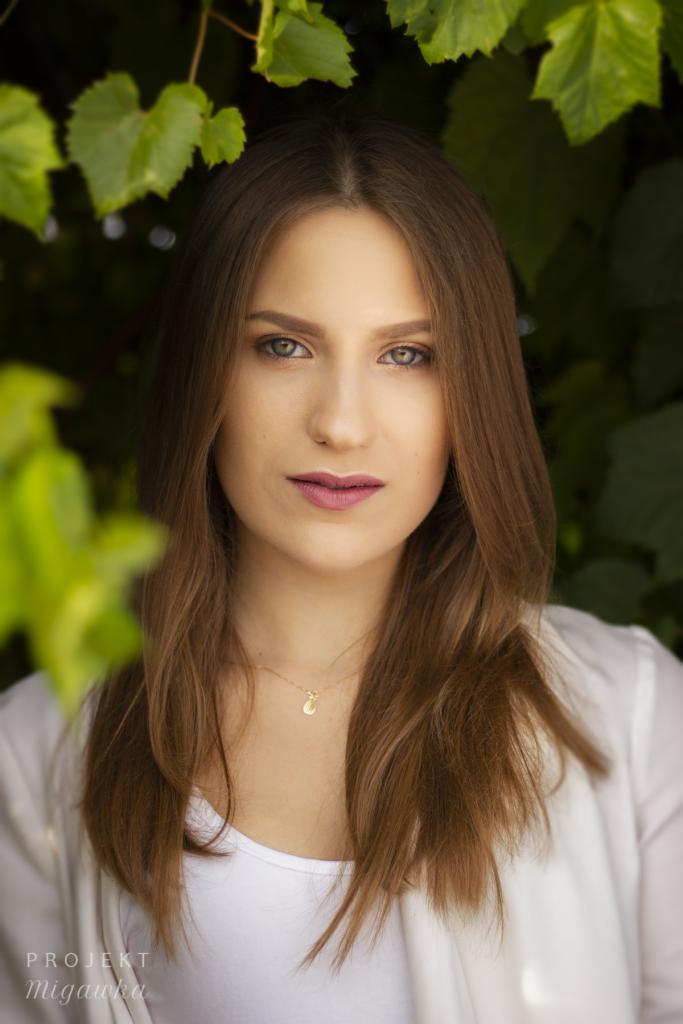 Portret młodej kobiety z długimi brązowymi włosami, zielonymi oczami i delikatnym makijażem, na tle zielonych liści winogron, z widocznym złotym naszyjnikiem z zawieszką.