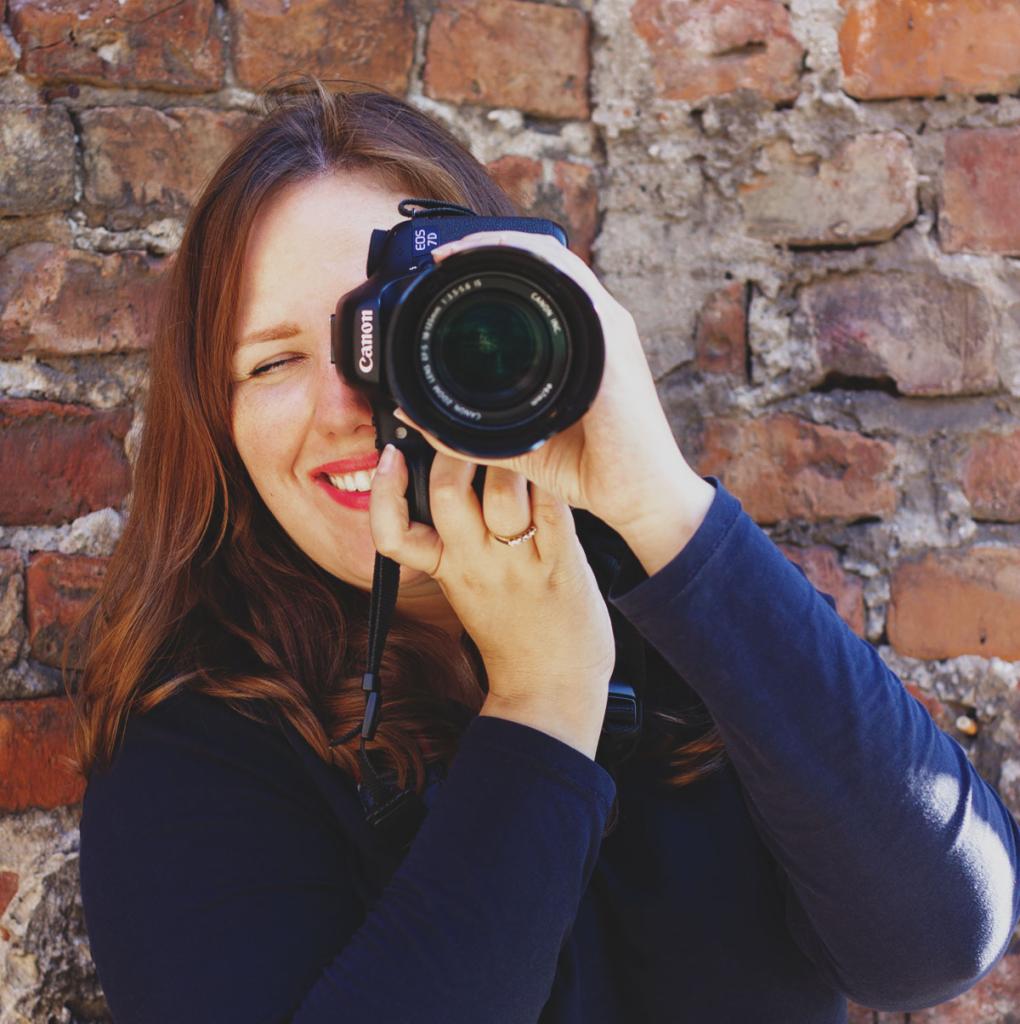 Uśmiechnięta kobieta z aparatem Canon EOS 7D, przymykająca oko, na tle ceglanej ściany.