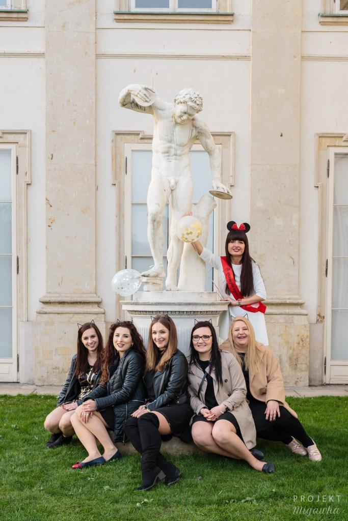 Grupa sześciu kobiet w różnym wieku, siedząca na trawie przed rzeźbą dyskobola i architekturą klasycystyczną, jedna kobieta w białej sukience z szarfą i uszami Myszki Miki trzyma balon, pozostałe...