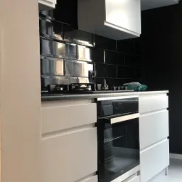 Nowoczesna kuchnia z błyszczącymi czarnymi płytkami na ścianie, białymi szafkami i piekarnikiem Beko. Na blacie widoczne elementy kuchenki gazowej i akcesoria.