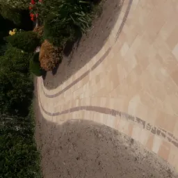 Zakrzywiony chodnik z beżowej kostki brukowej z ciemnobrązowymi obramowaniami, obok rabaty z zielonymi krzewami i czerwonymi kwiatami.