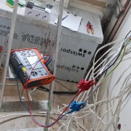 Przenośny miernik elektryczny na drabinie, z przewodami podłączonymi do wiązki kabli w trakcie prac instalacyjnych w pomieszczeniu w stanie surowym.
