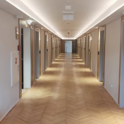 Lux-Instal - Długi korytarz z parkietem ułożonym w jodełkę, oświetlony listwami LED pod sufitem. Widoczne drzwi po obu stronach i czujnik dymu na suficie.