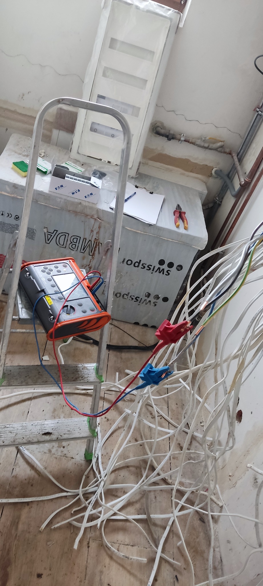 Przenośny miernik elektryczny na drabinie, z przewodami podłączonymi do wiązki kabli w trakcie prac instalacyjnych w pomieszczeniu w stanie surowym.