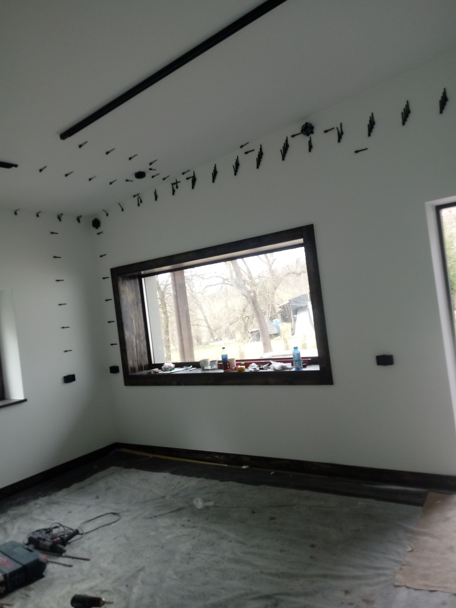 Wnętrze pokoju w trakcie instalacji systemu szynoprzewodów oświetleniowych. Widoczne czarne szyny i punkty montażowe na suficie i ścianach, przygotowane pod przyszłe lampy. Na podłodze folia...