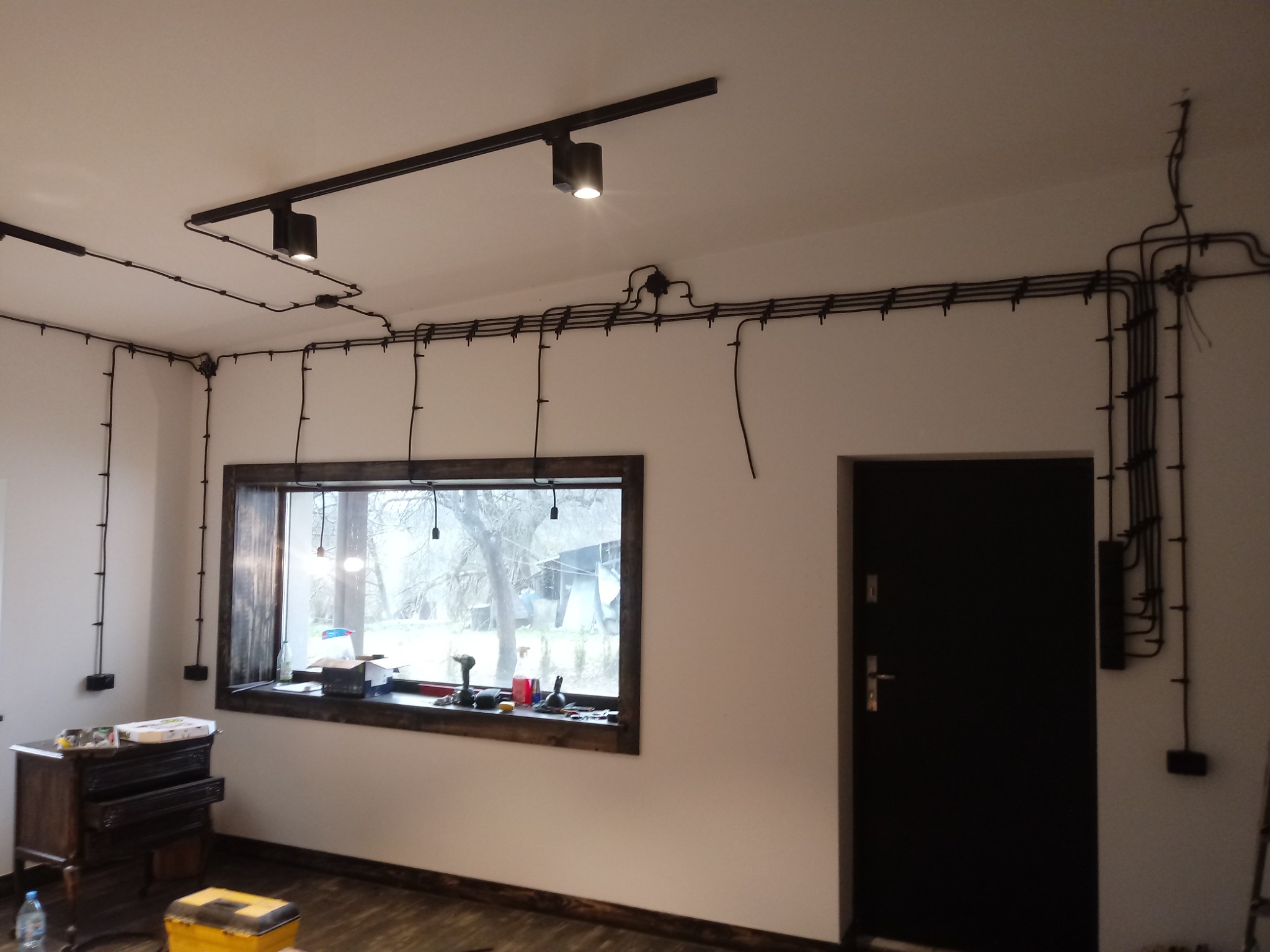 Wnętrze pokoju z widoczną instalacją elektryczną natynkową w czarnych rurkach, lampy sufitowe szynowe włączone, okno z drewnianą ramą, skrzynia narzędziowa i komoda w tle.