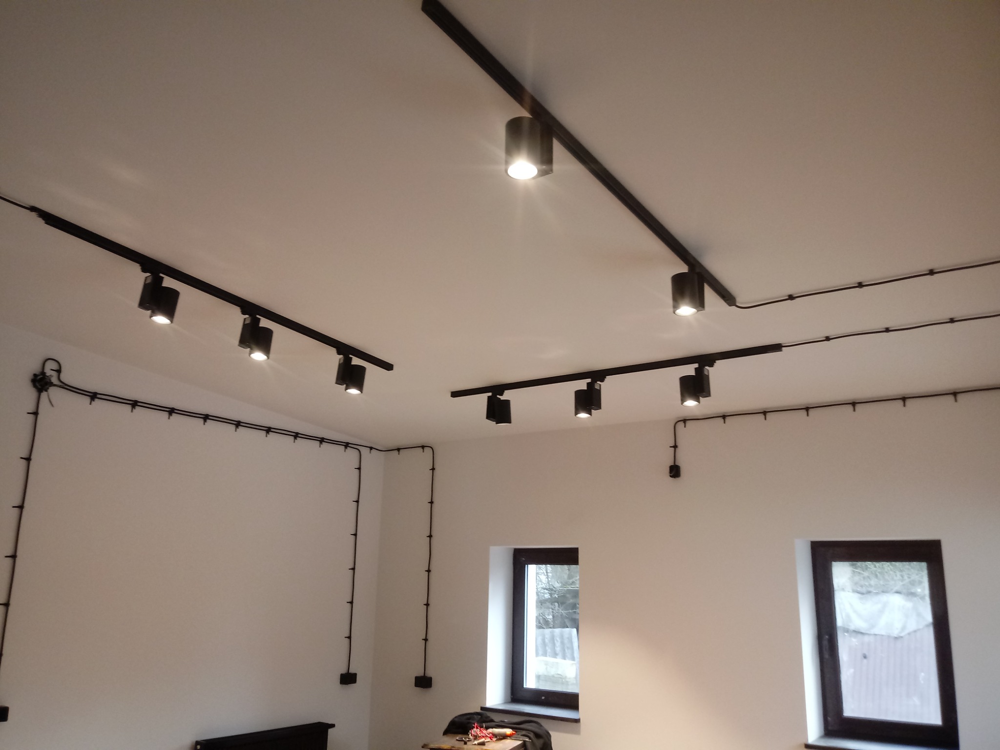 Czarna szyna z reflektorami sufitowymi w pomieszczeniu w Tucznie, widoczne poprowadzone kable elektryczne po ścianach, dwa okna z ciemnymi ramami.