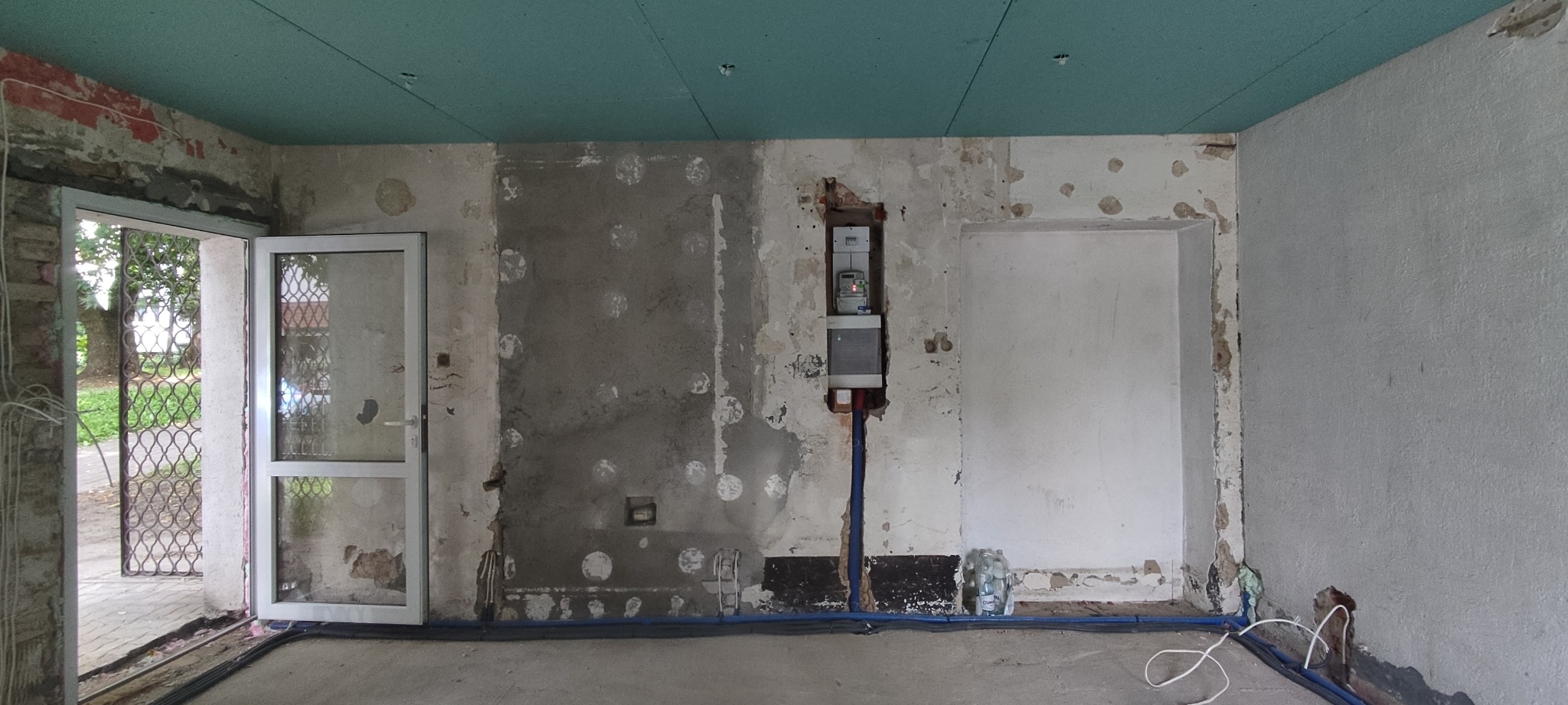 Wnętrze w trakcie remontu z odsłoniętą instalacją elektryczną, widoczna skrzynka z licznikiem, nieotynkowane ściany z pozostałościami farby, niebieskie rurki instalacyjne na podłodze i przy ścianie...
