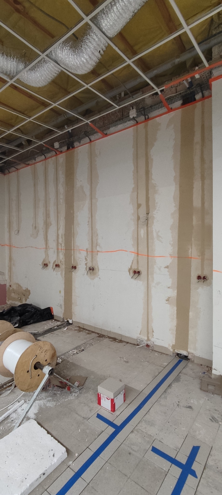Wnętrze w trakcie prac remontowych: widoczne przewody elektryczne wystające ze ściany, rolki kabli na podłodze oraz zabezpieczenia podłoża taśmą malarską.