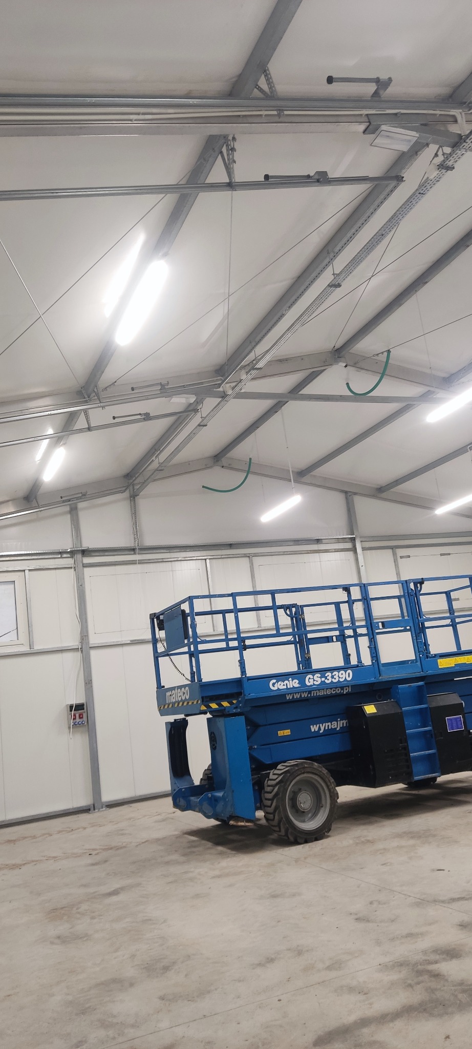 Wnętrze hali z widoczną podnośnikiem koszowym Genie GS-3390, zainstalowane lampy LED na metalowej konstrukcji sufitu.