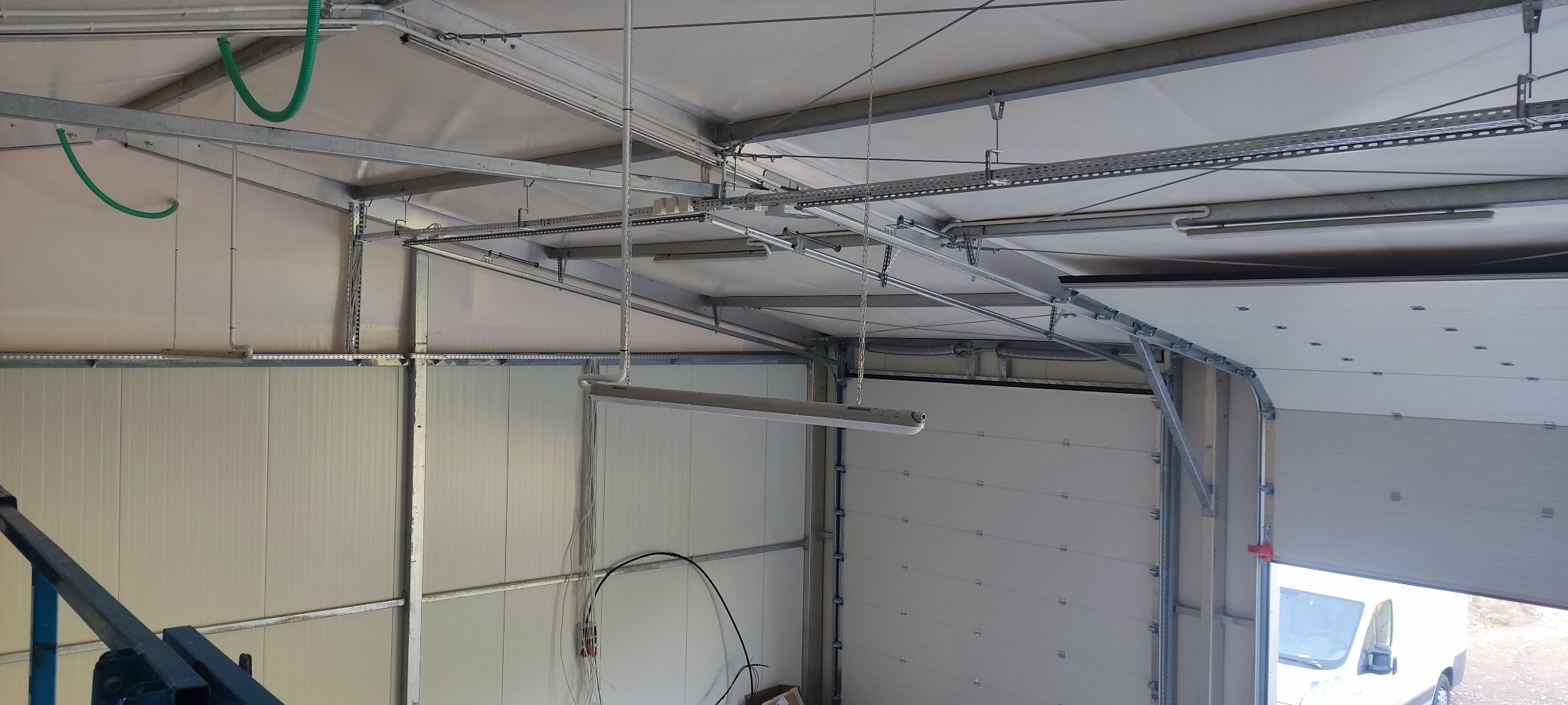 Wnętrze hali magazynowej z widocznymi elementami konstrukcyjnymi, systemem oświetlenia w postaci lamp wiszących na łańcuchach, oraz instalacją elektryczną biegnącą pod sufitem w korytkach kablowych...