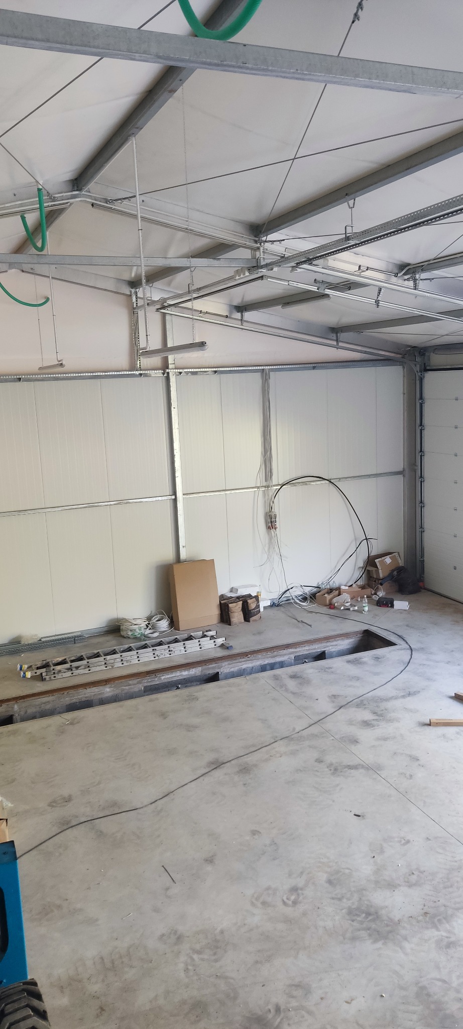 Wnętrze hali produkcyjnej w trakcie prac instalacyjnych, widoczne przewody elektryczne, drabina i materiały budowlane leżące na posadzce, oświetlenie sufitowe w trakcie montażu.