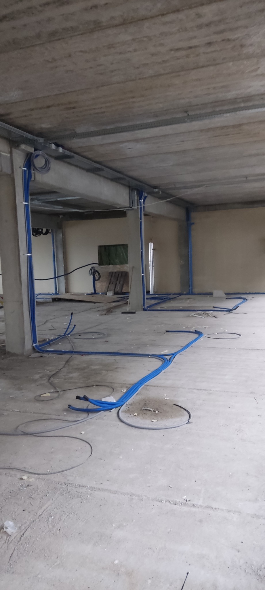 Niebieskie rury instalacji elektrycznej prowadzone po podłodze i pod sufitem w surowym wnętrzu budynku w trakcie budowy. Widoczne kable i elementy mocujące.