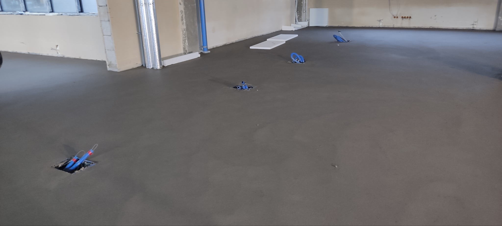 Wylana posadzka z wystającymi niebieskimi rurami instalacji elektrycznej w nowym budynku. Widoczne otwory w podłodze i ściany w stanie surowym.