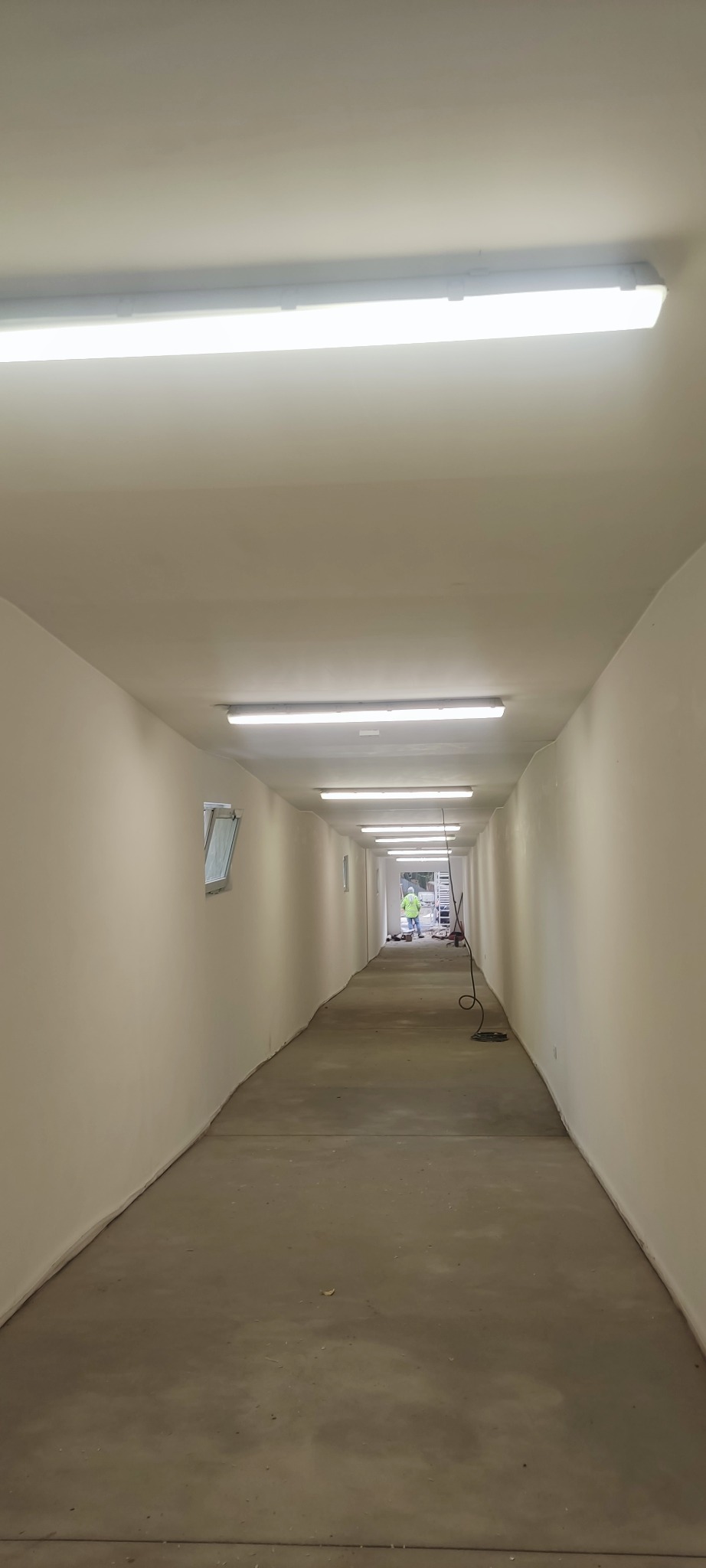 Długi korytarz z rzędem lamp fluorescencyjnych na suficie. W tle widoczna osoba w odblaskowej kamizelce i drabina. Ujęcie z perspektywy podłogi.