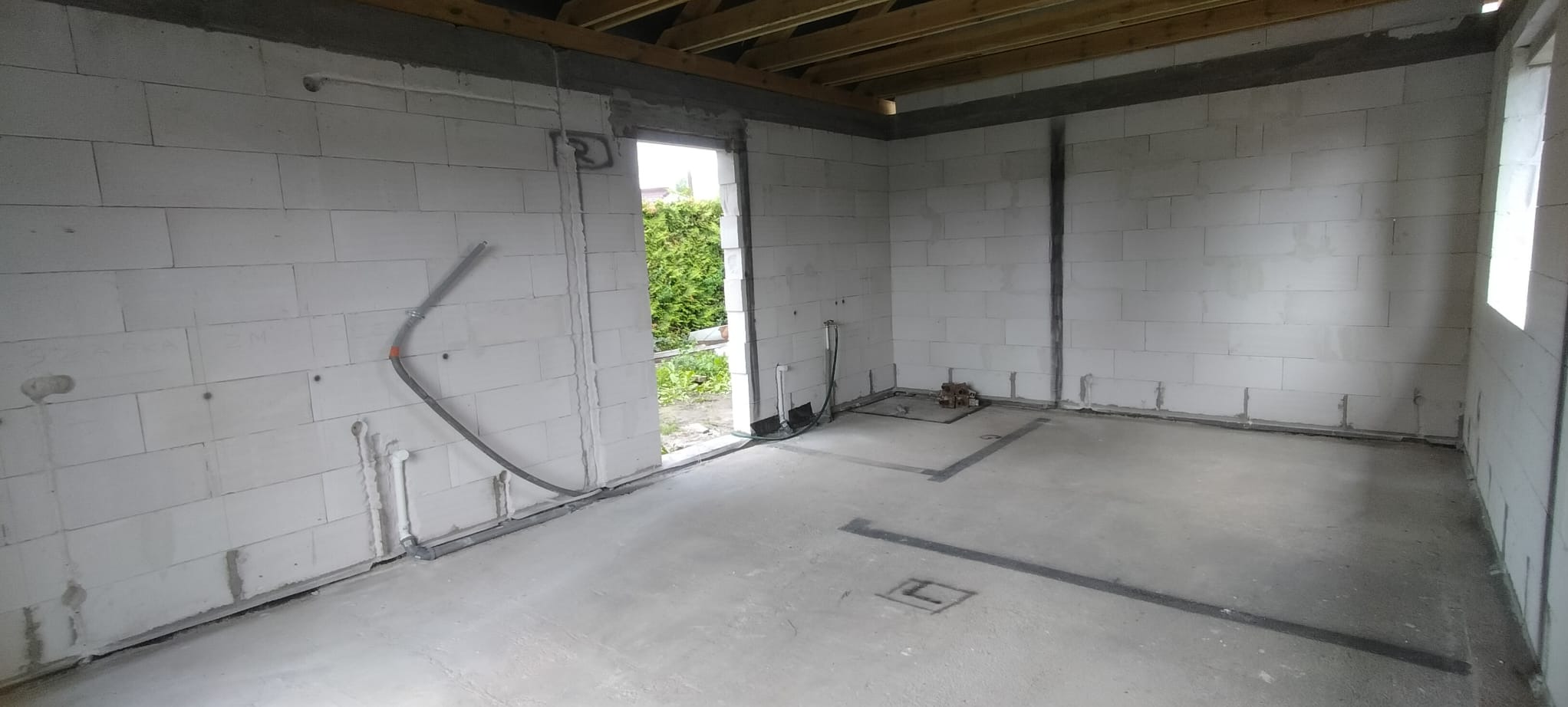 Surowy stan wnętrza z instalacją elektryczną w ścianach z betonu komórkowego. Widoczne kable i rury, przygotowane do dalszych prac wykończeniowych. Otwór drzwiowy z widokiem na zieleń.