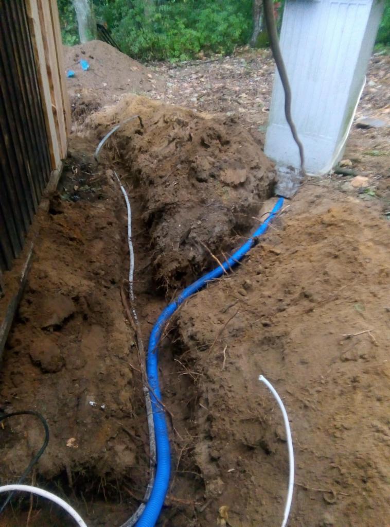 Wykop w ziemi z ułożonymi przewodami elektrycznymi w peszlach, widoczne kable i fragment budynku z drewnianą elewacją.