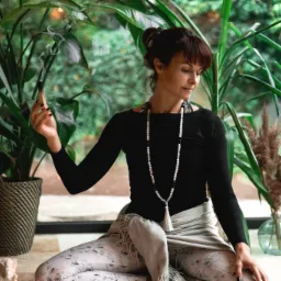 Kobieta w pozycji lotosu, trzymająca tlące się Palo Santo na tle bujnej zieleni, z długim naszyjnikiem z koralików i szalem na kolanach.