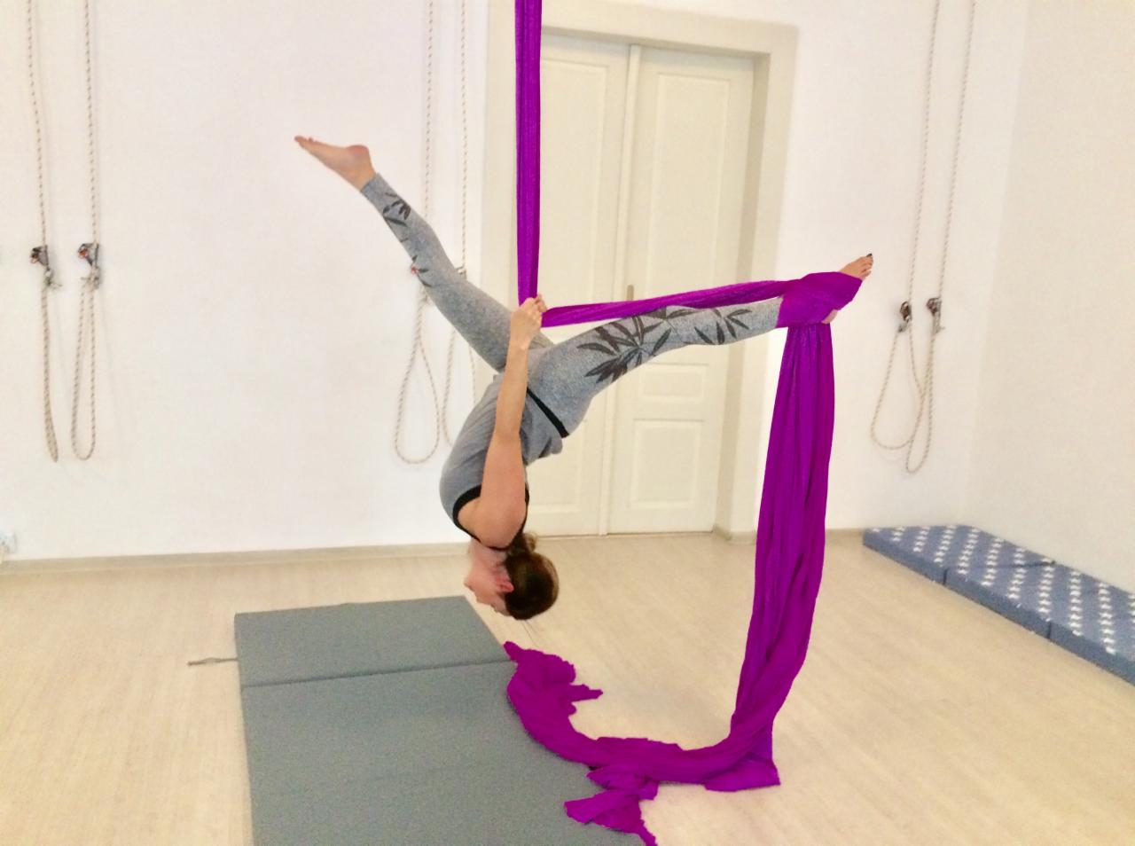 Kobieta w szarym stroju z motywem liści wykonuje pozycję odwróconą na fioletowej chuście do aerial silks w jasnym studio z drewnianą podłogą i białymi ścianami.