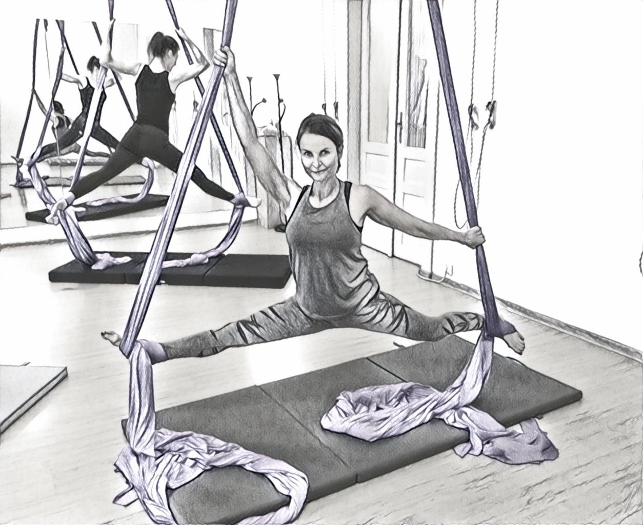 Kobieta uśmiecha się, wykonując szpagat w powietrzu z pomocą jedwabnych chust podwieszonych do sufitu, widoczne maty do ćwiczeń i lustra w tle.