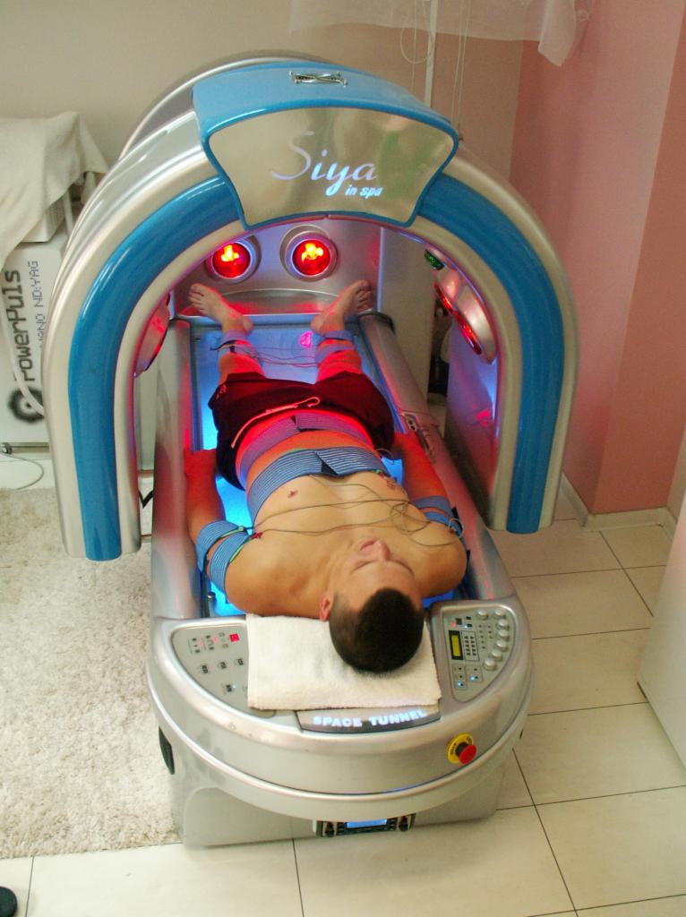 Mężczyzna leżący w kapsule SPA z niebieskim i czerwonym światłem, podłączony do elektrod, na białym ręczniku, z widocznym panelem sterowania kapsuły.