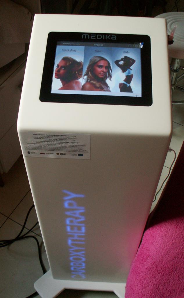 Urządzenie Medika Carboxytherapy z ekranem dotykowym prezentującym zabiegi na skórę głowy, twarz i ciało. Widoczna naklejka informacyjna i przewody zasilające.