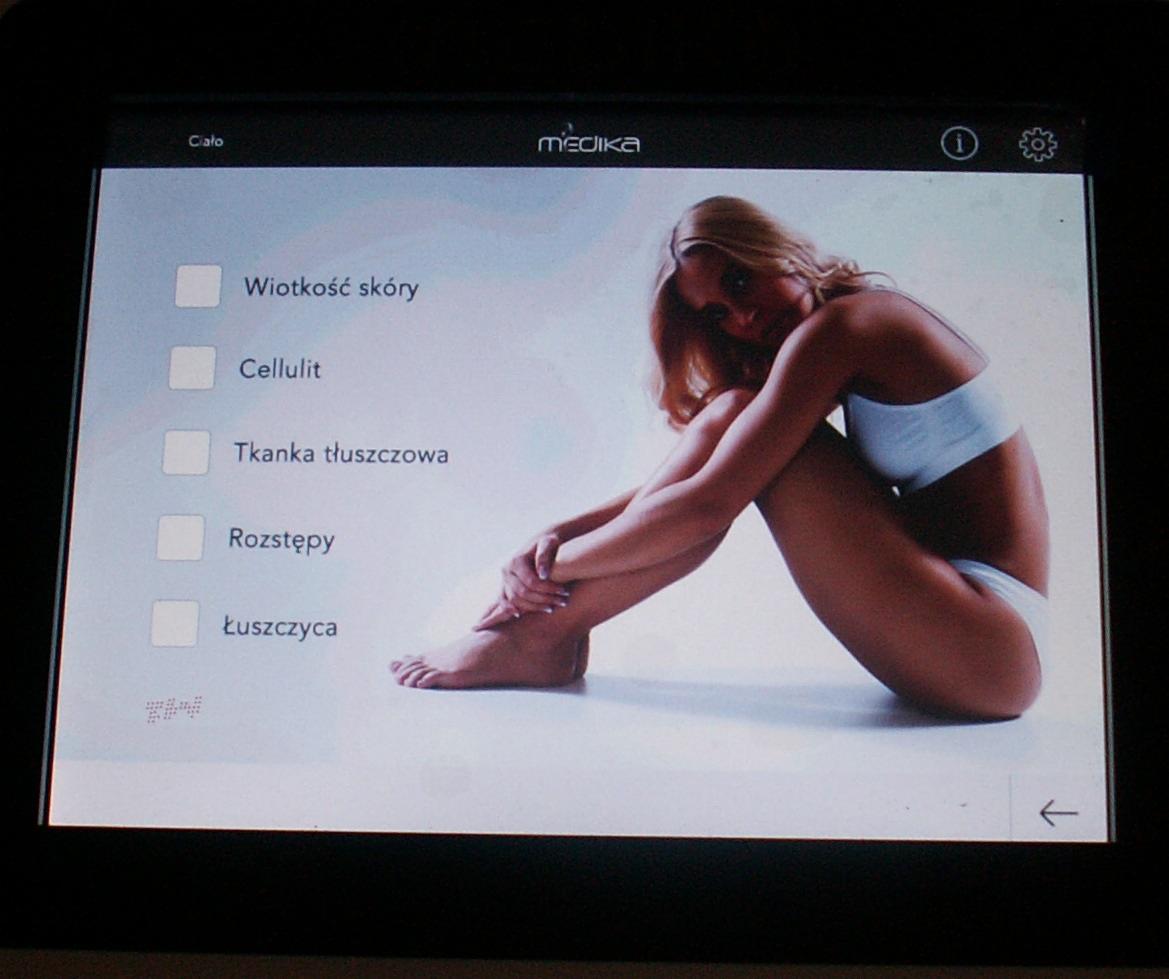 Ekran urządzenia z listą problemów skórnych (wiotkość, cellulit, tkanka tłuszczowa, rozstępy, łuszczyca) i zdjęciem kobiety w bieliźnie.