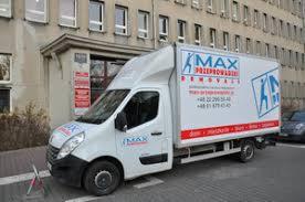 Biała furgonetka transportowa z napisem 'MAX PRZEPROWADZKI REMOVALS' zaparkowana na ulicy w mieście, na tle budynku mieszkalnego z licznymi oknami. Widoczny fragment chodnika i jezdni.