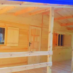 Jasny, drewniany domek z niebieskim dachem, widok z zewnątrz na werandę i las w tle.