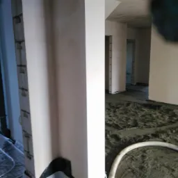 Wylewanie posadzki jastrychowej z widocznym systemem ogrzewania podłogowego, rurą doprowadzającą mieszankę i surowymi ścianami w tle.