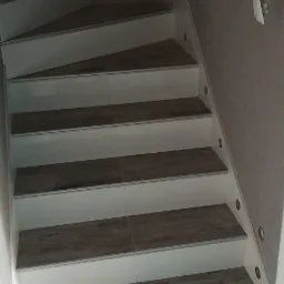 Wykończone schody z drewnopodobnymi płytkami na stopniach i białymi podstopnicami, z oświetleniem punktowym w ścianie, widoczna instalacja elektryczna w tle.