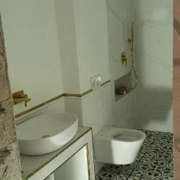 Wnętrze łazienki w Sokołowie Podlaskim w trakcie wykańczania, widoczne białe sanitariaty, umywalka nablatowa, toaleta, kabina prysznicowa z złotą armaturą oraz czarno-biała wzorzysta podłoga.