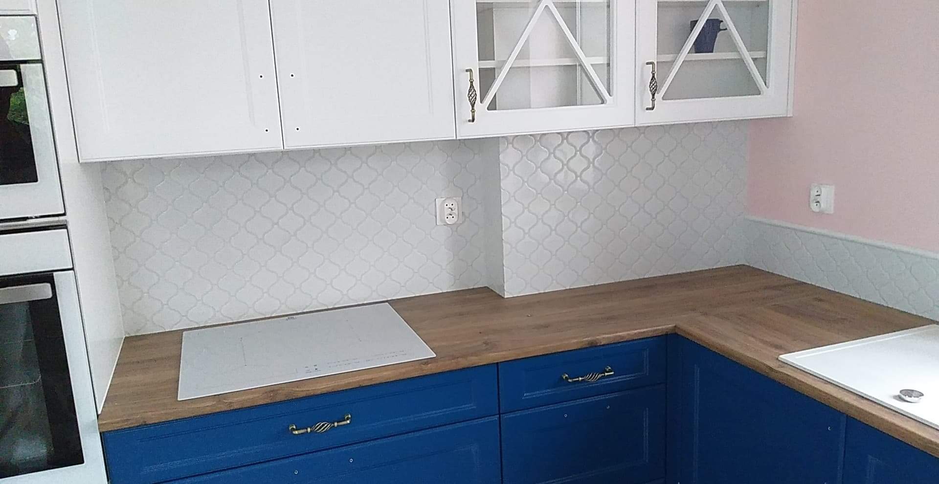 Wykończona kuchnia z białymi szafkami górnymi z przeszklonymi drzwiczkami, niebieskimi szafkami dolnymi z ozdobnymi uchwytami, drewnianym blatem i białymi płytkami w marokański wzór na ścianie.
