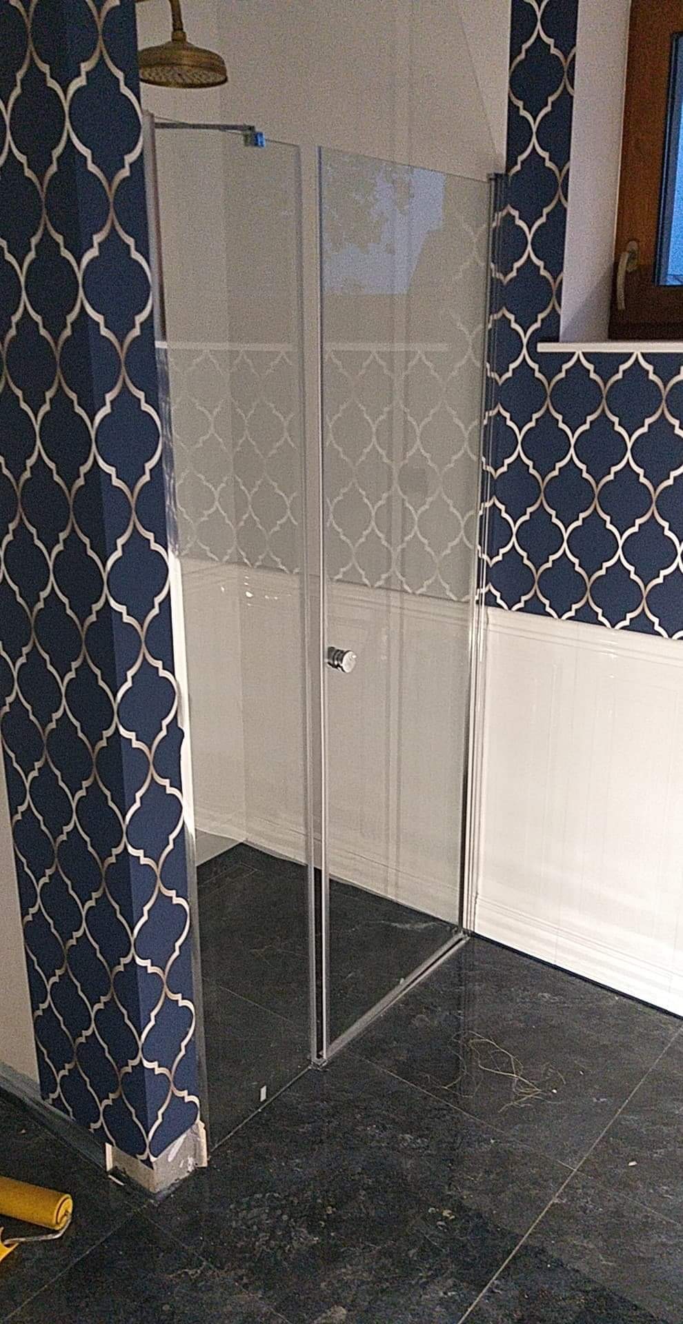 Nowoczesna kabina prysznicowa ze szklanymi ścianami, zamontowana na ciemnej, marmurkowej podłodze. Ściany wykończone białą boazerią i niebieską tapetą w złoty wzór. Mosiężna deszczownica widoczna...