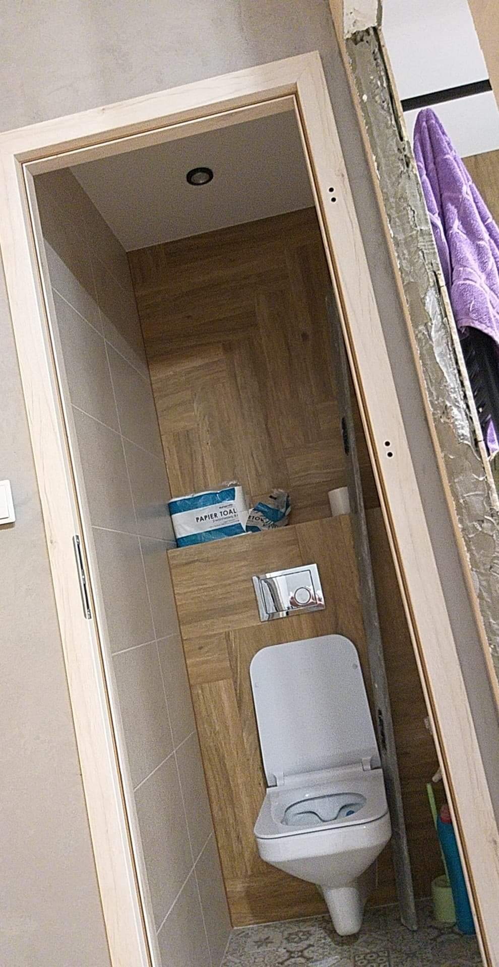 Wnętrze łazienki w trakcie remontu, widoczna toaleta podwieszana, płytki imitujące drewno na ścianach i podłodze, niedokończone obramowanie drzwi, ręcznik i środki czystości w rogu.