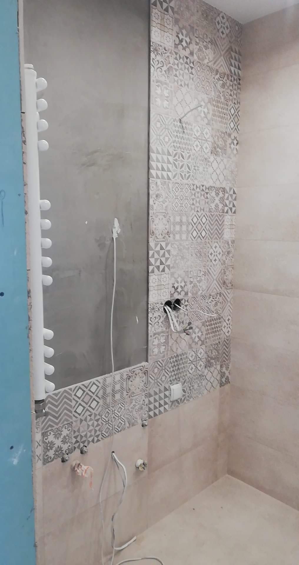 W trakcie prac wykończeniowych w łazience: fragment ściany z położonymi wzorzystymi płytkami ceramicznymi, obok ściana przygotowana pod płytki, widoczne wystające kable i rury, grzejnik drabinkowy.
