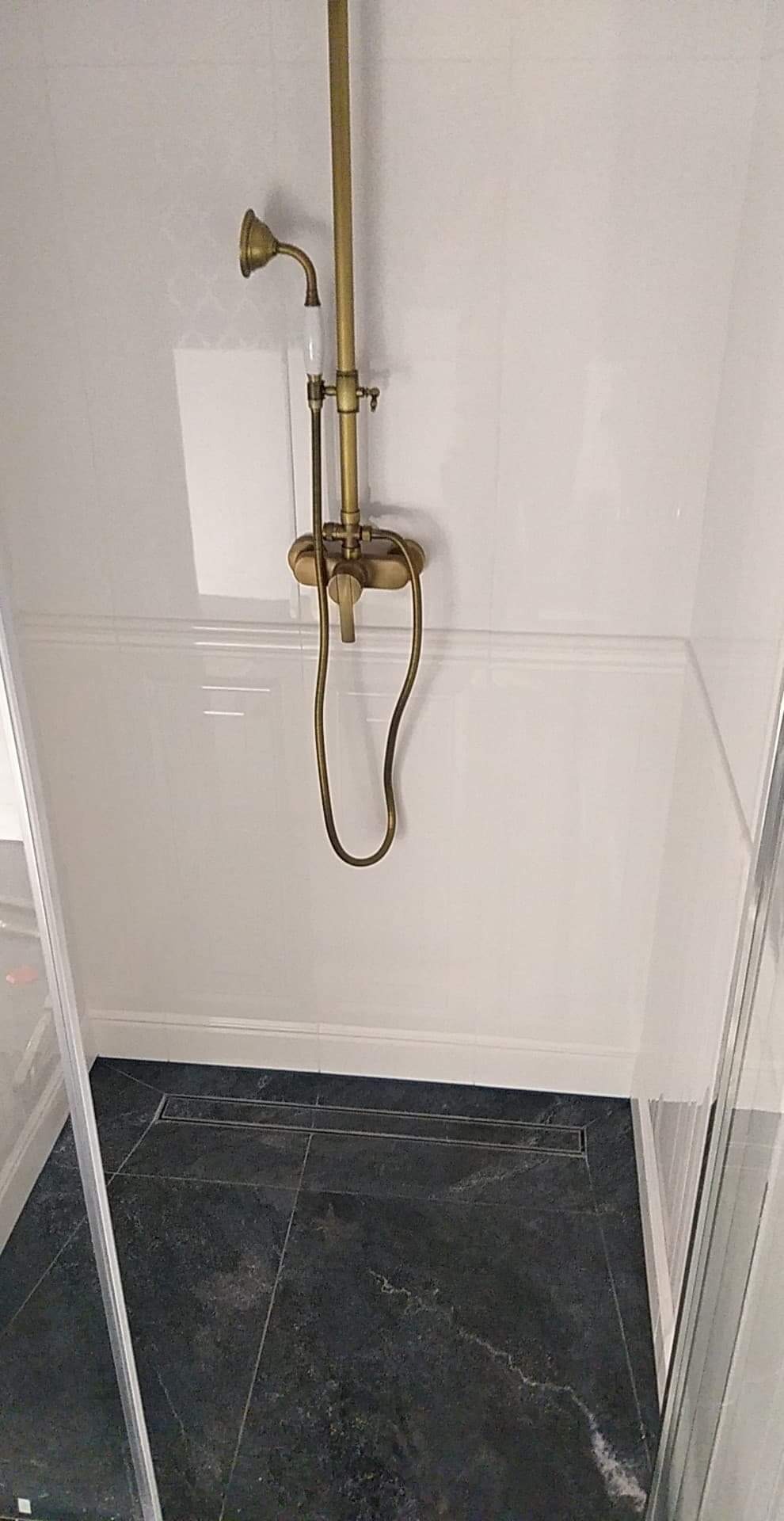 Kabina prysznicowa z armaturą w stylu retro w kolorze starego złota i odpływem liniowym w posadzce wyłożonej ciemnymi płytkami imitującymi marmur.