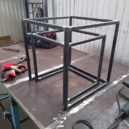 Metalowa konstrukcja w kształcie trzech zagnieżdżonych sześcianów, spawana na stole roboczym, widoczne ślady spawania, rękawice robocze i narzędzia w tle.