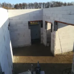 Wnętrze budynku w trakcie murowania ścian z bloczków białych, widoczne otwory drzwiowe z nadprożami, na podłodze narzędzia i resztki materiałów budowlanych.