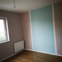 Pokój po remoncie: ściany pomalowane na brązowo i błękitno, drewniana podłoga, biały grzejnik pod oknem, wystający kabel ze ściany.